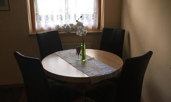 A small dining area with a round table and four black chairs. On the table is a glass bottle with a flower and a table runner.
