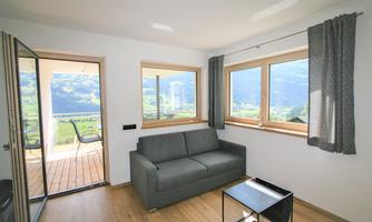 A cozy living room with a gray sofa and large windows. The view of the mountains and the terrace creates a bright, inviting atmosphere.