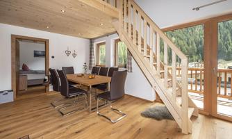 A modern living room with a wooden dining area. The large window front offers a view of the greenery and the balcony.