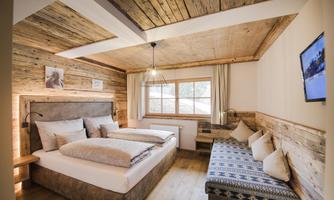 A cozy bedroom with wooden walls and a large window. There is a comfortable bed and a seating area with cushions.