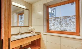 A modern bathroom with a wood vanity and a large mirror. The window has a decorative curtain and lets in natural light.