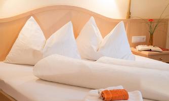A cozy bedroom with a large bed and six decorative pillows. On the bedspread lies a towel along with a small flower and a book.
