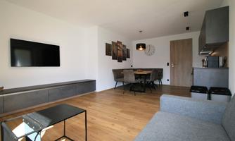 A modern living room with a gray sofa and a television on the wall. The room has wooden flooring and a dining table with several chairs.