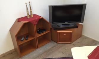 A small living area with a television and a wooden chest of drawers. Two shelves are located next to it, one with remote controls and a small device.
