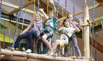 Four children are smiling and happily posing on a climbing structure. They are wearing safety harnesses and standing on wooden platforms.