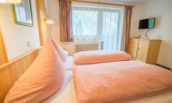 A cozy hotel room with two beds and pink pillows. Daylight streams through the windows, and a TV is mounted on the wall.