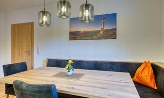 A cozy dining area with a wooden table and blue chairs. A picture hangs on the wall, and there are modern lamps as well as a small floral decoration on the table.