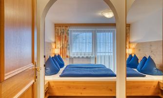 A cozy bedroom with wooden decor and blue pillows. There are large windows that let in natural light.