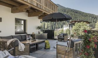 A cozy terrace with modern furniture and a sun umbrella. Green mountains can be seen in the background.