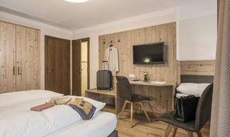 A modern hotel room with two single beds and wood-paneled walls. There is a table with a chair, a television, and a suitcase next to the bed.