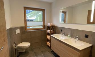 A modern bathroom with double sinks and wooden shelves. Large windows provide plenty of daylight.