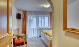 A cozy hotel room with a double bed, a chair, and a small table. The window is covered with curtains, and the room has soft colors.