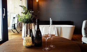 A stylish table with a flower arrangement, a bottle of sparkling wine, and two glasses. In the background, a modern bathtub and a cozy room are visible.