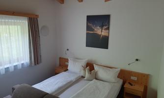 A cozy bedroom with two single beds and warm wooden walls. A picture hangs on the wall and large windows let in a lot of light.
