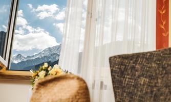 A view from a window overlooking the mountains and a clear sky. In the foreground, a comfortable armchair and a small floral decoration can be seen.