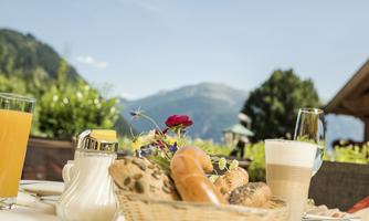 A breakfast table with fresh rolls, jam, and drinks. In the background, green mountains and a blue sky can be seen.