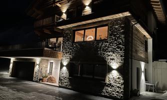 A modern wooden house at night, illuminated by warm lights. The stone structure stands out against the dark background.