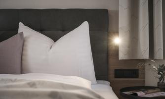 A cozy bed with large, white pillows and a blanket. Next to the bed is a small table with a plant and a lamp.
