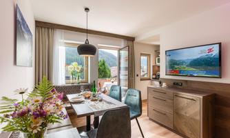 A modern apartment with a dining area and a television. Large windows offer a view of nature.