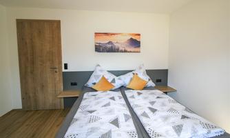 A cozy bedroom with two single beds, gray bedding, and colorful pillows. On the wall hangs a picture of a mountain landscape.