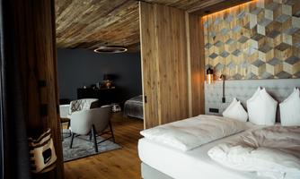 A modern bedroom with a cozy bed and wooden ceiling. In the background, there is another room with stylish furniture.