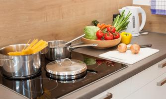A modern kitchen with pots on the stove and fresh vegetables on the countertop. Next to the ingredients are also some onions and a kettle.