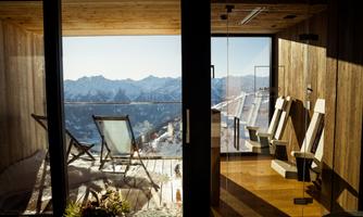 A modern room with large windows and a view of snow-capped mountains. Outside, lounge chairs are placed on a terrace that invites relaxation.