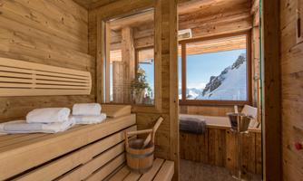 A modern sauna with wood paneling and a stunning view of snow-covered mountains. Towels and a wooden container for water are also available.