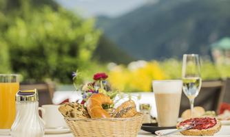 An appealing breakfast buffet with fresh rolls, jam, and various drinks. In the background, green mountains and flowers can be seen.