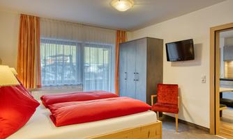 A cozy bedroom with a double bed, red pillows, and a modern television. Large windows let in plenty of daylight, and there is a comfortable chair as well as a wardrobe.