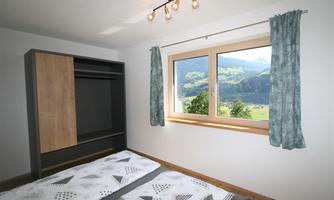 A simple bedroom with a wardrobe and a large window. A green landscape can be seen through the window.