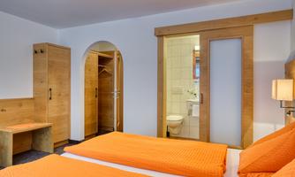 A cozy bedroom with two orange beds and a wooden table. In the background, a bathroom with a white door is visible.
