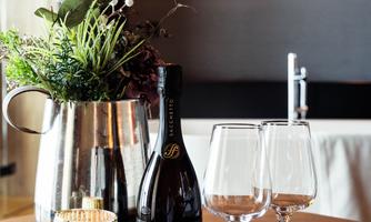 A bottle of sparkling wine is on a table, surrounded by two glasses and a modern botanical decoration. In the background, soft lighting elements can be seen.