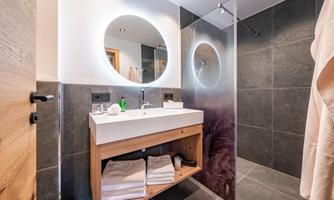A modern bathroom with a large mirror light and a minimalist wooden sink. The walls are in dark stone, and towels are neatly stored.