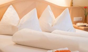 A cozy bedroom with three large pillows and fresh towels. On the nightstand, there is a small flower.