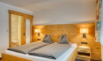 A cozy bedroom with a double bed and two large pillows. The walls are adorned with wood paneling and elegant lighting is installed.