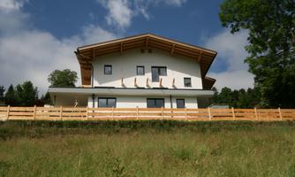 A modern house with a large roof and wooden cladding. It stands in a green meadow under a blue sky.