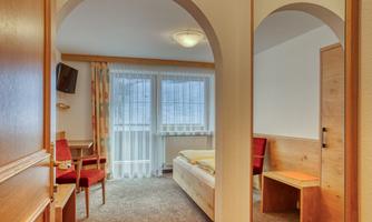 A simple room with two red chairs, a table, and a bed. Large windows provide plenty of light and a cozy atmosphere.