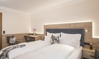 A modern bedroom with a large double bed and wooden paneling. The room is bright and inviting, with a desk and a small flower decoration.