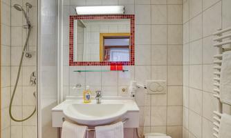 A modern bathroom with a shower, sink, and mirror. White tiles and towels create a bright, clean atmosphere.