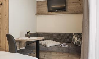 A cozy living area with a corner bench and a table. A television is mounted on the wall and a small plant is on the table.