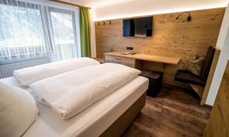 A modern room with a double bed and large windows. There is a desk and a television on the wall.