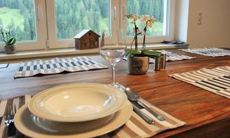 A beautifully set table with plates, cutlery, and a wine glass. In the background, there are windows overlooking a green forest landscape.