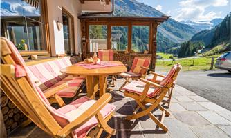 A cozy outdoor area with wooden furniture and a round table. In the background, green meadows and impressive mountains can be seen.