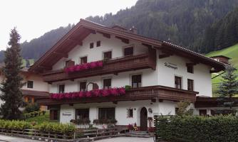 A charming alpine house with a balcony adorned with colorful flowers. In the background, green mountains and a cloudy sky can be seen.