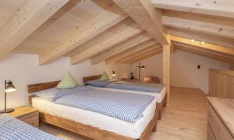 A cozy attic bedroom with three beds and wooden furniture. The wood ceiling and warm lighting create an inviting atmosphere.
