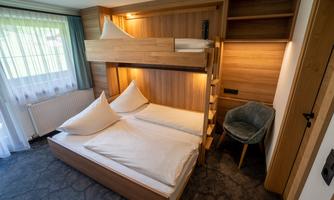 A modern hotel room with a bunk bed and a double bed. The wooden paneling and large windows create a cozy atmosphere.