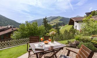 A terrace with a wooden table and chairs, surrounded by green landscapes and mountains. In the background, typical alpine houses can be seen.