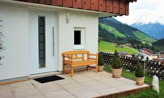 A cozy terrace with a wooden bench and two plants. In the background, green hills and snow-capped mountains can be seen.