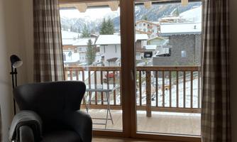 A cozy room with a black armchair and a footstool. Through the windows, one can see snow-covered mountains and a quiet village.